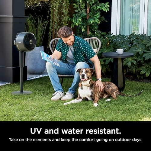 Man sitting outdoors with a dog, reading a magazine, next to a floor fan.